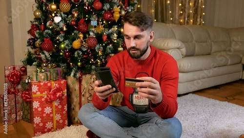 Young caucasian man red sweater buy gifts online for xmas in front of christmas tree using his smartphone. High quality 4k footage
