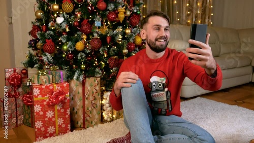 Young caucasian man red sweater making videocall online in front of christmas tree using his smartphone. High quality 4k footage