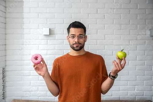 Young student man doubts what to choose healthy food or sweets junk unhealthy food holding green apple and donuts in hands standing at home in her living room. Hard choice concept eating disorder photo
