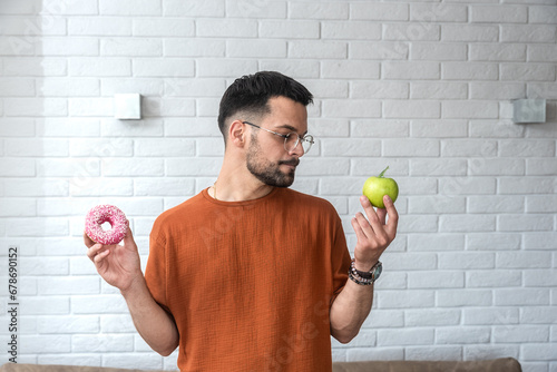 Young student man doubts what to choose healthy food or sweets junk unhealthy food holding green apple and donuts in hands standing at home in her living room. Hard choice concept eating disorder photo