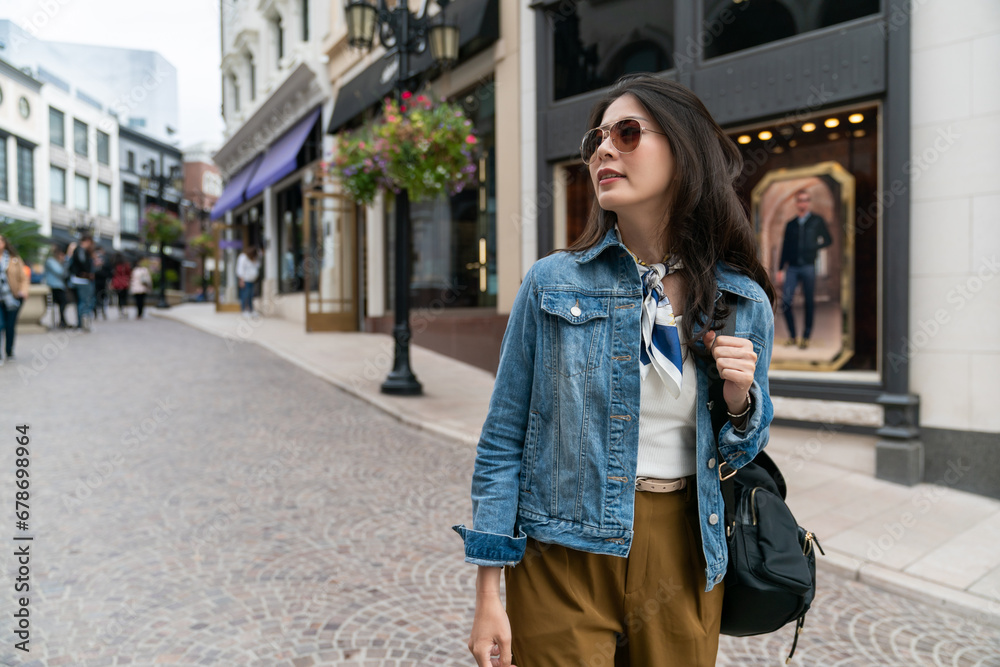 Fototapeta premium Asian woman with black hair wearing sunglasses walking on city streets while doing shopping outdoors. Sales and discounts concept.
