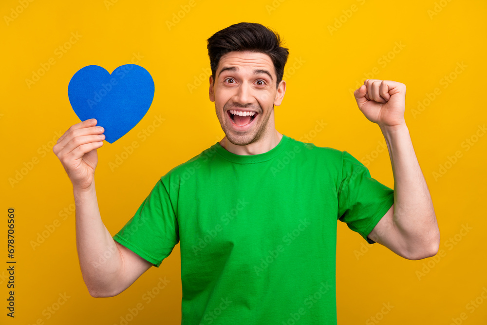 Photo portrait of nice young brunet male raise fist celebrate hold blue heart wear trendy green garment isolated on yellow color background Photo portrait of nice young brunet male raise fist celebrate hold blue heart wear trendy green garment isolated on yellow color background