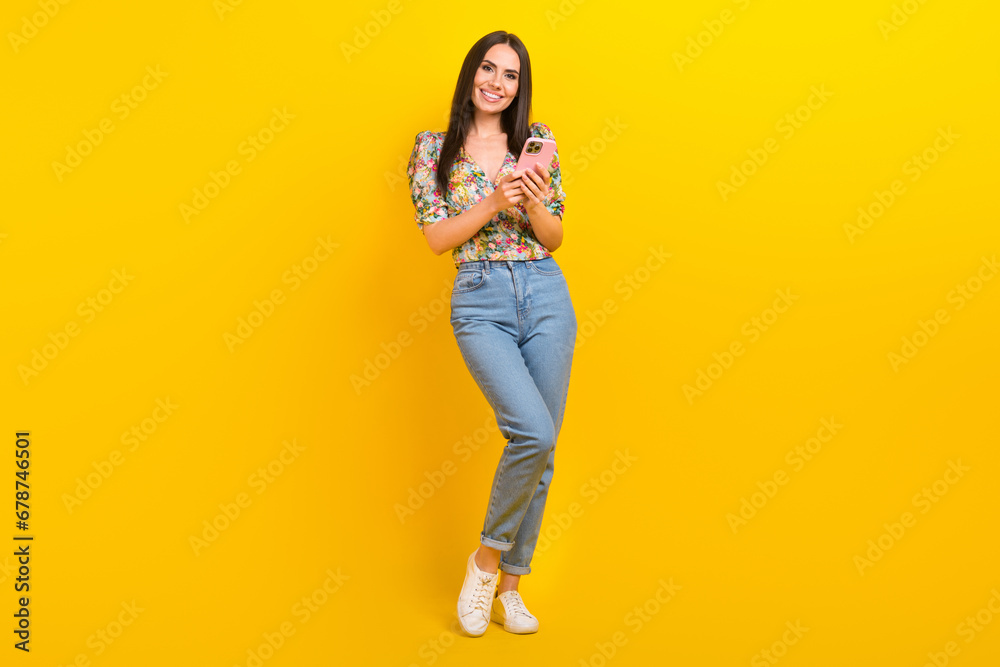 Full length photo of satisfied cute girl wear flower print blouse jeans hold smartphone read email isolated on yellow color background Full length photo of satisfied cute girl wear flower print blouse jeans hold smartphone read email isolated on yellow color background