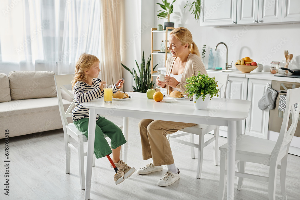 preschooler girl with prosthesis leg having breakfast with happy blonde mother in modern kitchen