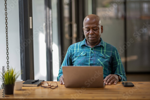 A black senior man in casual clothes is focusing on a laptop screen, concentrating and thinking seriously.