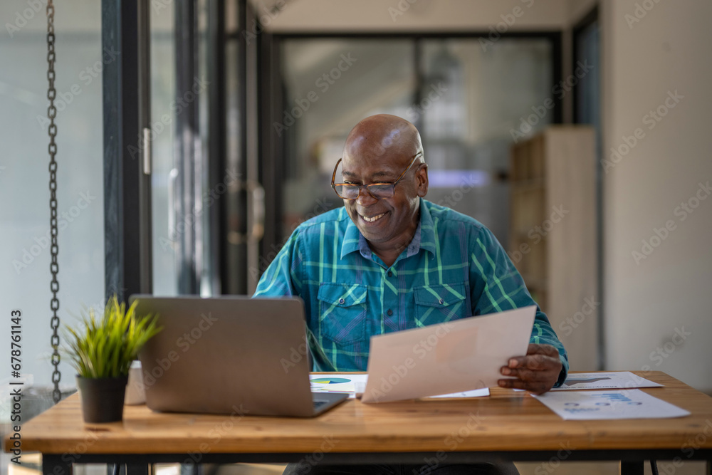 A senior black man in casual clothes is busy at work but happy with the work and paperwork in front of him.