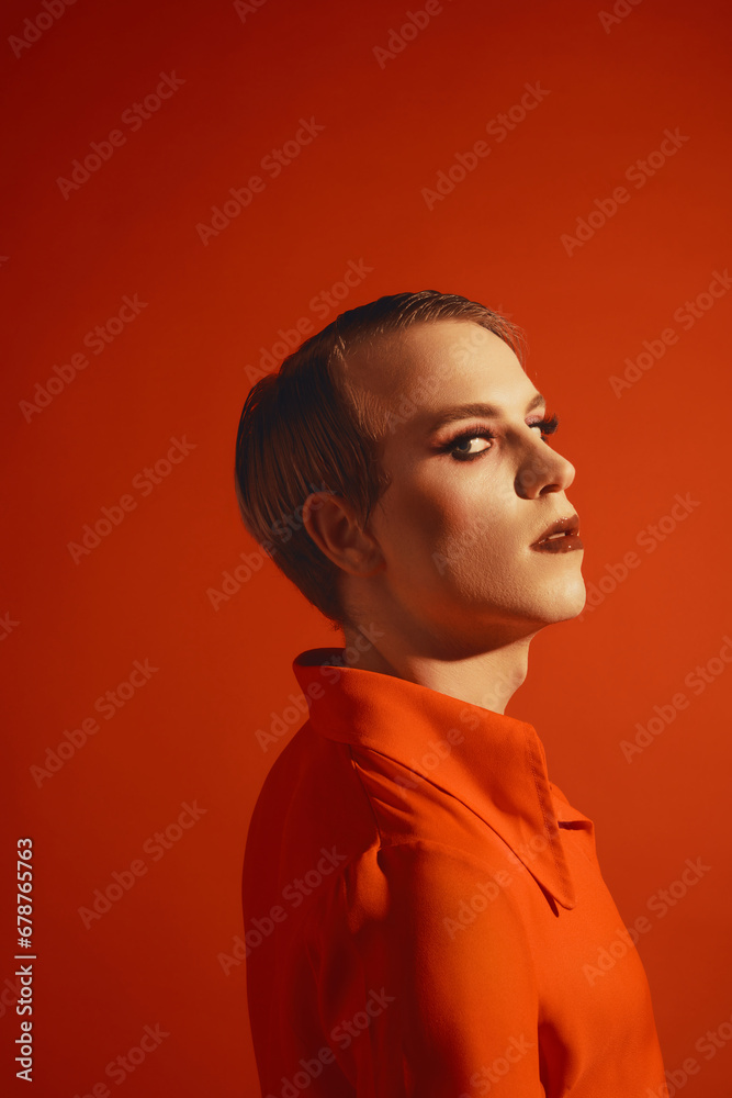 Portrait of young guy in bright jacket, makeup with colorful eyes and lashes posing against red studio background. Concept of male makeup, fashion, lgbtq community, self-identity, acceptance Portrait of young guy in bright jacket, makeup with colorful eyes and lashes posing against red studio background. Concept of male makeup, fashion, lgbtq community, self-identity, acceptance