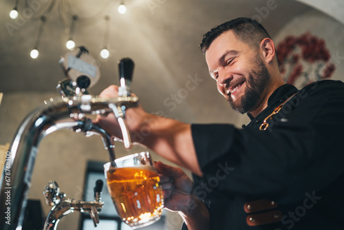 Smiling stylish bearded barman dressed black uniform with an apron tapping fresh lager beer into glass mug at bar counter. Successful people, beer consumption, beverages industry concept image