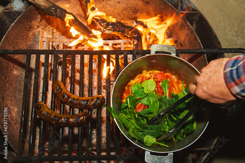 A hand with tongs cooking food in an outdoor grill over a hot campfire outdoors, seen from above photo