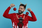 Happy young man in Christmas sweater and reindeer headband holding festive baubles on light blue background