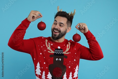 Happy young man in Christmas sweater and reindeer headband holding festive baubles on light blue background