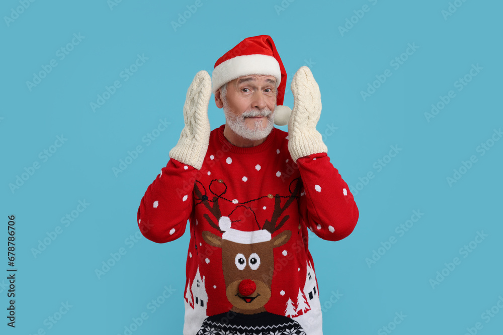 Senior man in Christmas sweater, Santa hat and knitted mittens on light blue background