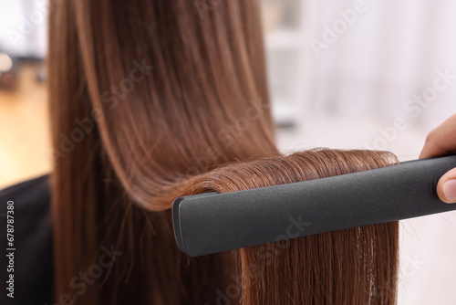 Hairdresser straightening woman's hair with flat iron indoors, closeup photo