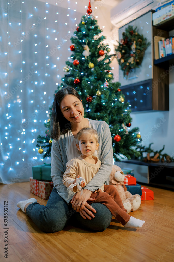 Smiling mother with a little girl on her knees sits near a decorated Christmas tree on the floor