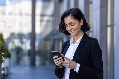 A young beautiful business woman is walking outside the office building, a woman in business clothes is smiling, using an application on the phone, browsing the Internet, typing a message