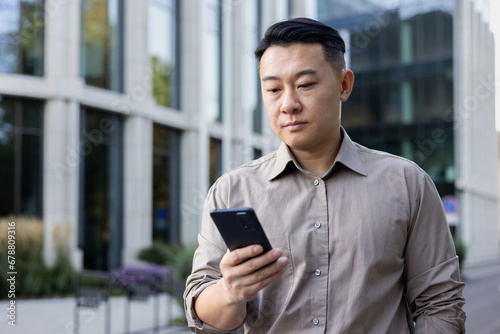 Close-up photo of a young Asian male businessman standing outside an office center with a phone in his hands, waiting for an appointment, making a call, typing a message