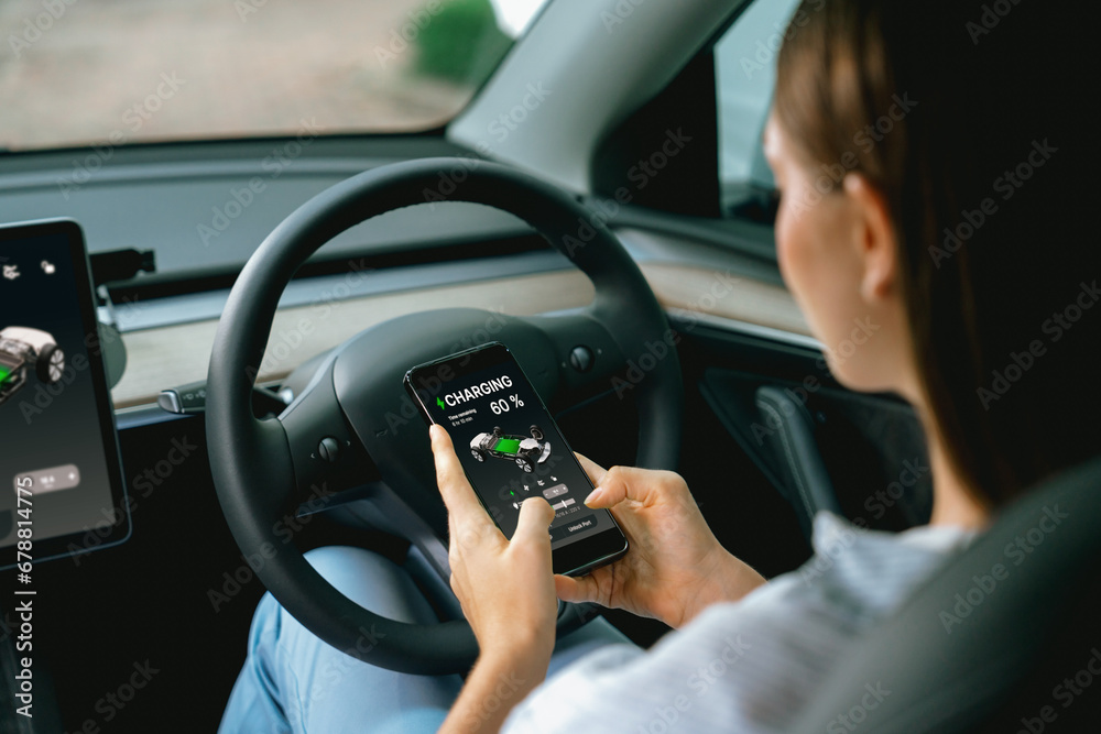 Young woman inside the vehicle, checking EV vehicle's application on battery recharging electricity status display on smartphone screen in modern EV car on her road trip journey. Exalt