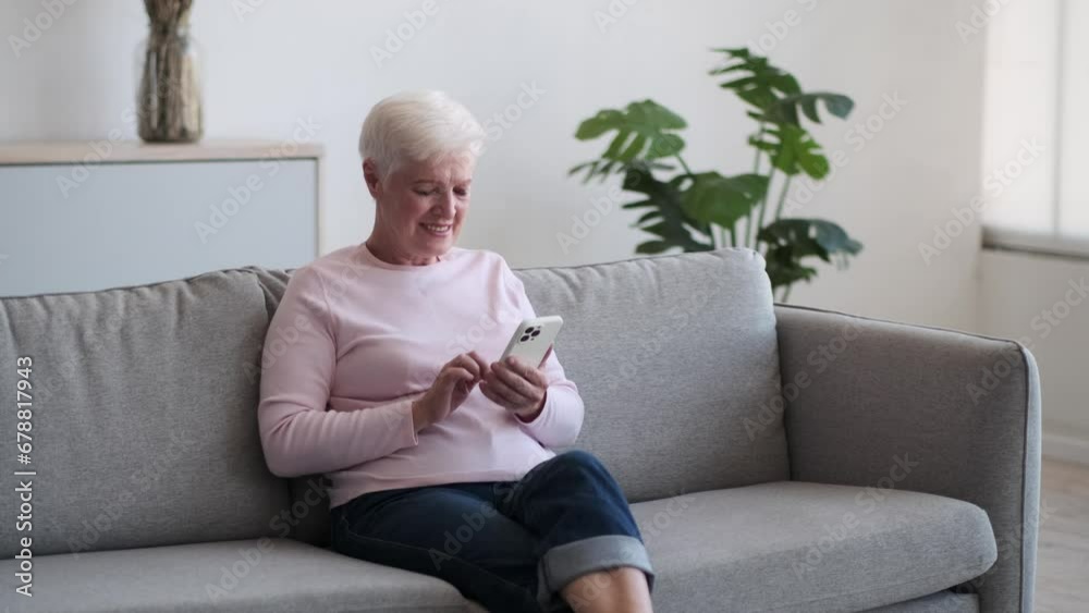 Wise elderly woman sits serenely on the couch, attention captivated by the screen of phone. Her fingers scroll with gentle precision, exploring the digital realm with a sense of curiosity. Wise elderly woman sits serenely on the couch, attention captivated by the screen of phone. Her fingers scroll with gentle precision, exploring the digital realm with a sense of curiosity.