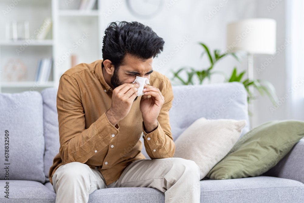 Sick man sitting at home on sofa in living room, sneezing has allergy and runny nose, Indian man with napkin has a cold. Sick man sitting at home on sofa in living room, sneezing has allergy and runny nose, Indian man with napkin has a cold.