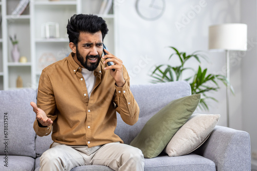 Angry angry man talking on the phone at home, dissatisfied customer sitting on the sofa in the living room yelling at the interlocutor.