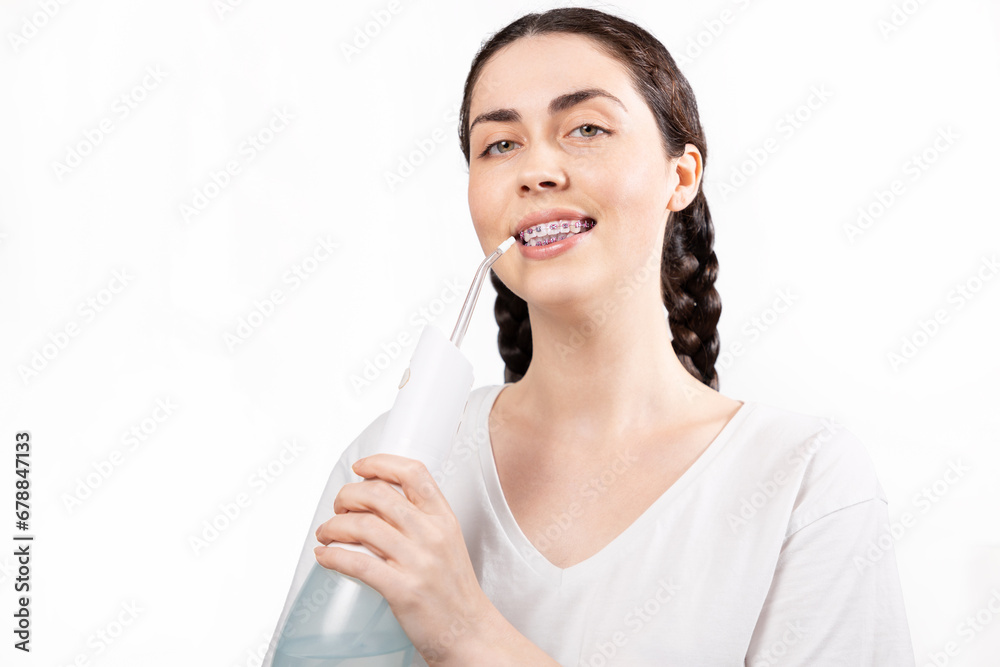 Portrait of young smiling woman with brackets on teeth cleans them using oral irrigator. White background. Concept of dental care during orthodontic treatment Portrait of young smiling woman with brackets on teeth cleans them using oral irrigator. White background. Concept of dental care during orthodontic treatment