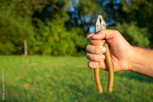 Close-up of a man's hand holding old dirty pliers against a green landscape. Sunlight, tools. Copy space photo