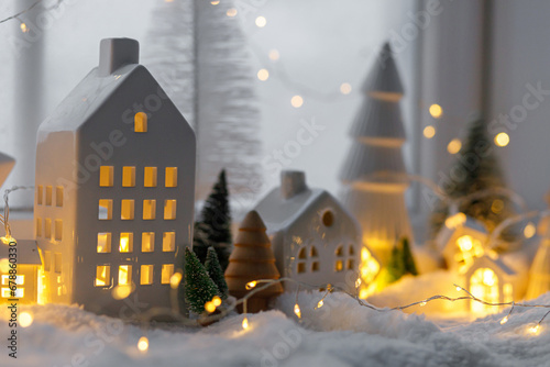 Atmospheric miniature winter village. Stylish little ceramic houses and christmas wooden trees on snow blanket with glowing lights in evening. Christmas modern background. Happy Holidays!