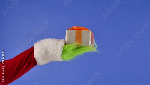 The Grinch's green haired hand holds out a white gift box with a orange bow on an isolated blue background. Gift kidnapper cosplay. Blue screen, chroma key. photo