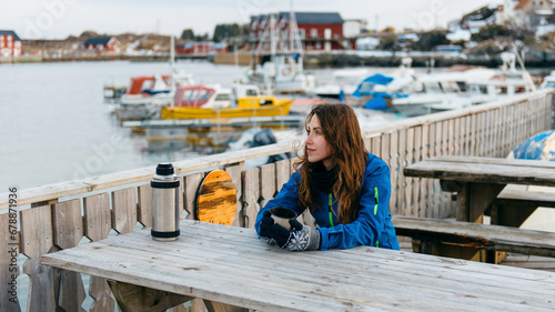 Happy smiling girl traveler drinks warm tea at the rustic wooden table outdoor near the traditional fishing houses Rorbu and fishing ships in Lofoten islands, Norway photo