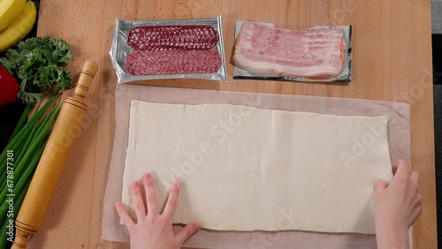 Puff pastry is laid out on a wooden table against a background of sausage, bicon, greens, female hands roll out rectangular dough with a wooden rolling pin. Top view, close-up photo