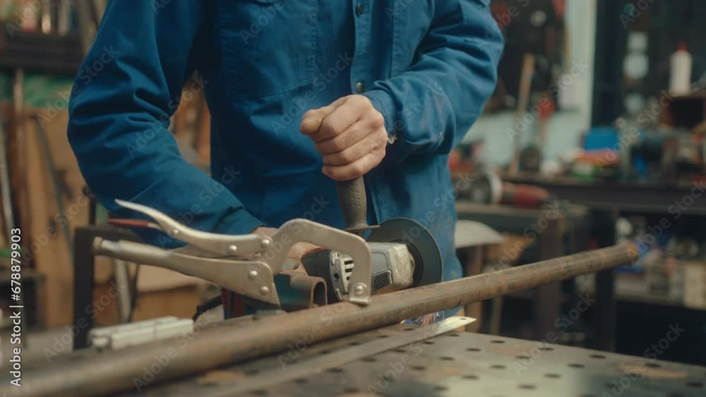 Professional metalsmith in protective eyewear standing by workbench and cutting pipe with disk grinder making sparks in the repair shop. Tilt-up shot Professional metalsmith in protective eyewear standing by workbench and cutting pipe with disk grinder making sparks in the repair shop. Tilt-up shot