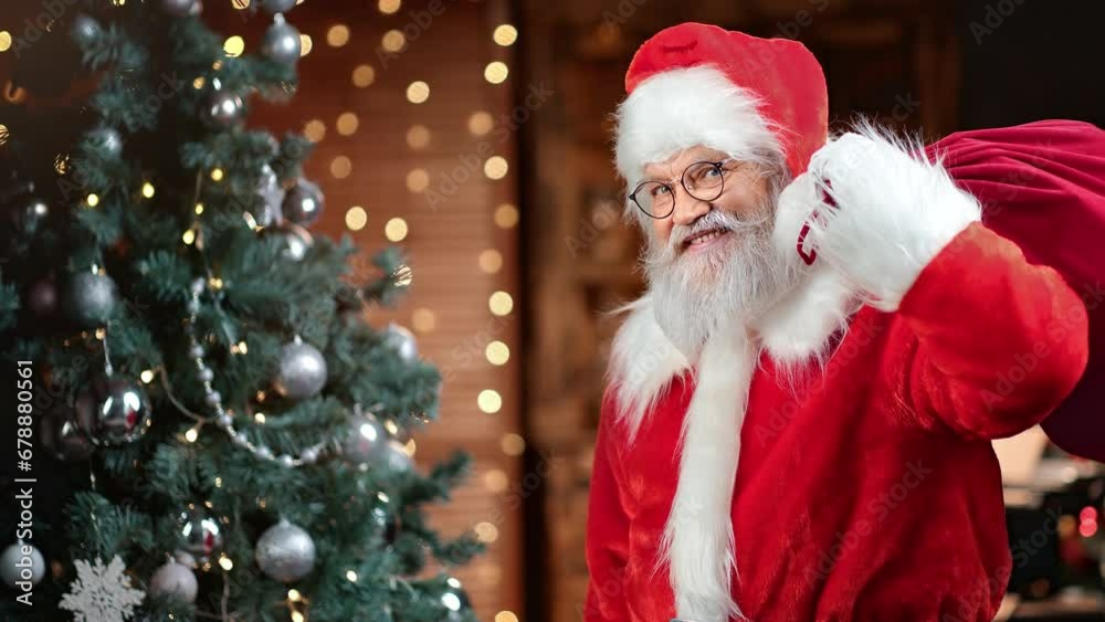 Smiling Santa Claus with red sack full of present posing near illuminated Christmas tree