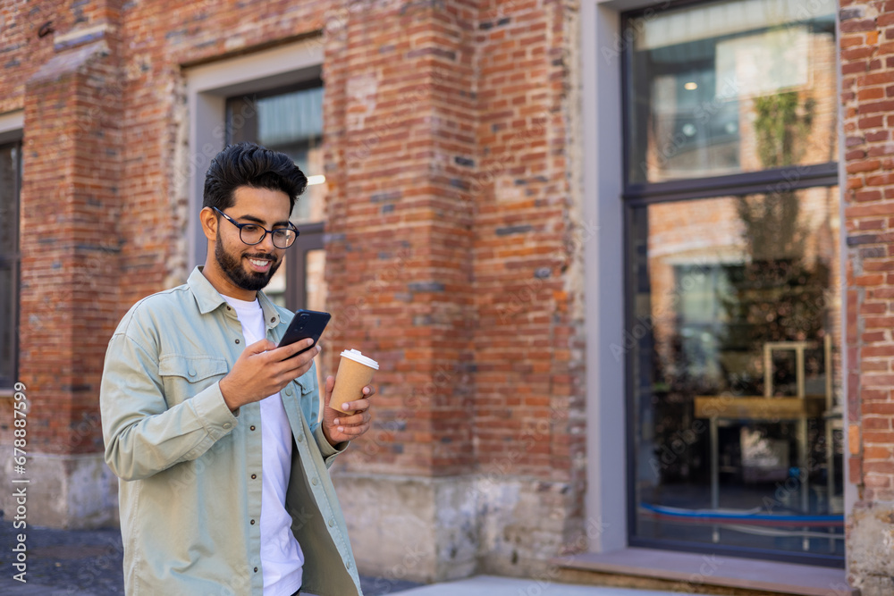 Young man walking in the city reading messages on the phone smiling, with a cup of hot drink, businessman entrepreneur programmer in casual clothes using an application on a smartphone Young man walking in the city reading messages on the phone smiling, with a cup of hot drink, businessman entrepreneur programmer in casual clothes using an application on a smartphone