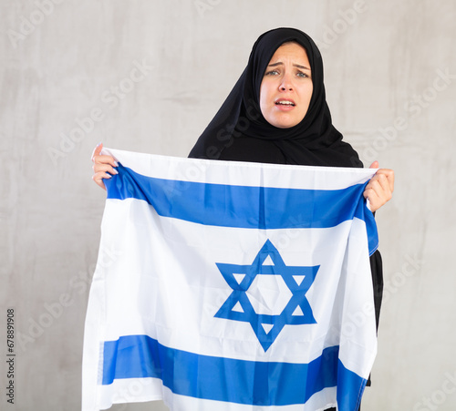 disappointed Muslim woman in traditional black hijab holds flag of Israel. Portrait of female Muslim with Israeli flag on gray background photo