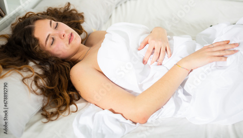 Attractive girl with long brown wavy hair sleeping on white bedlinen in her bed at home..