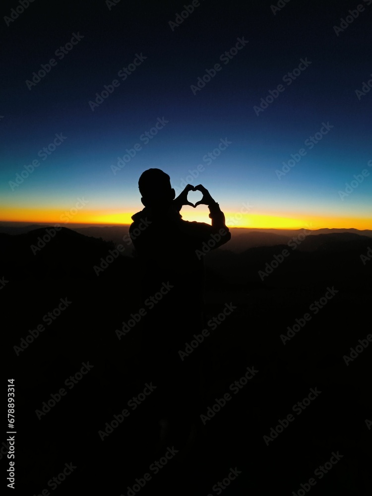 Vertical of a silhouette of a guy holding her hands in the shape of a heart against the sunset sky