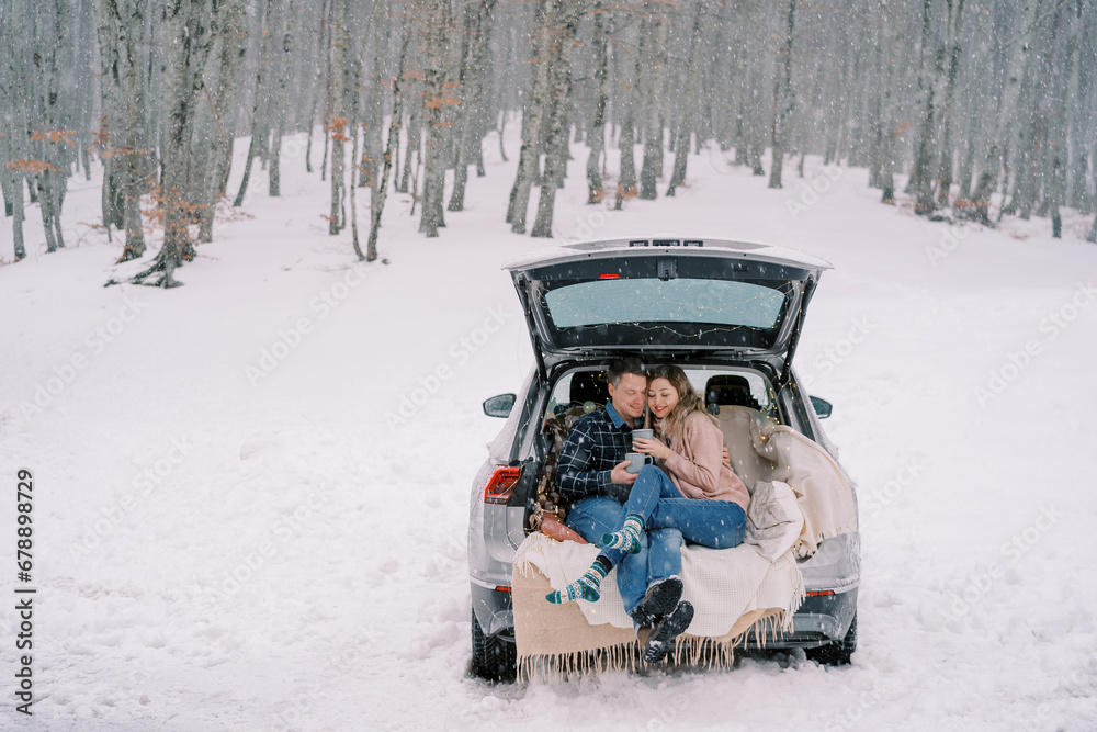 Couple with cups of coffee sits hugging in the trunk of a car on blankets under snowfall in the forest