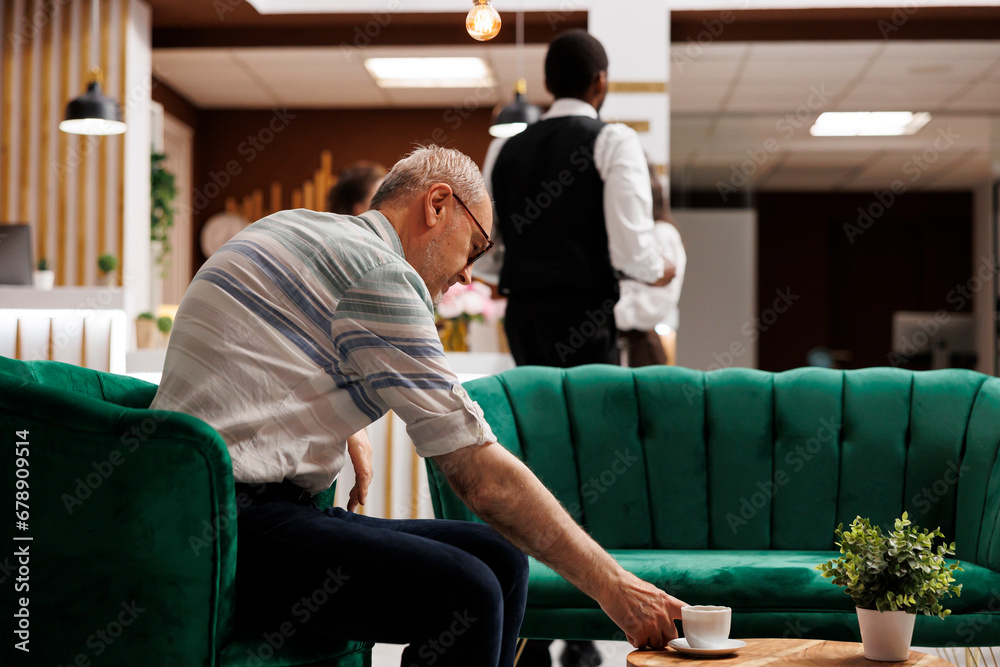 At elegant lounge area for check-in, elderly caucasian man sits on sofa reaching for cup of coffee on table. Senior guy in hotel lobby, waiting on the couch for the booking procedure. At elegant lounge area for check-in, elderly caucasian man sits on sofa reaching for cup of coffee on table. Senior guy in hotel lobby, waiting on the couch for the booking procedure.