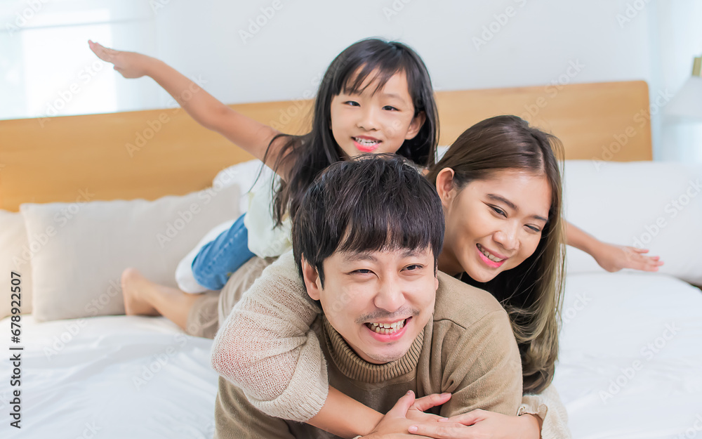 Three people of Asian family, father, mother, little daughter playing together with fun on bed in bedroom at cozy home, smiling with happiness. Kid, Housing, Healthcare Concept. Three people of Asian family, father, mother, little daughter playing together with fun on bed in bedroom at cozy home, smiling with happiness. Kid, Housing, Healthcare Concept.