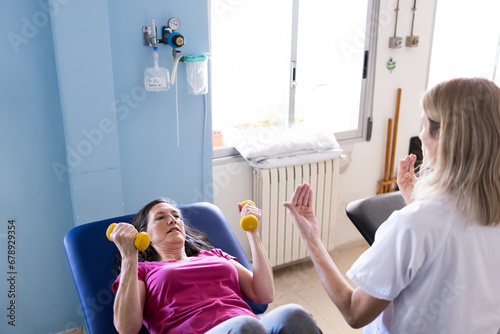 Focused photo of a middle aged patient with brown hair and a pink shirt while exercising with 1kg dumbbells and her physioterapist in front of her out of focus. photo