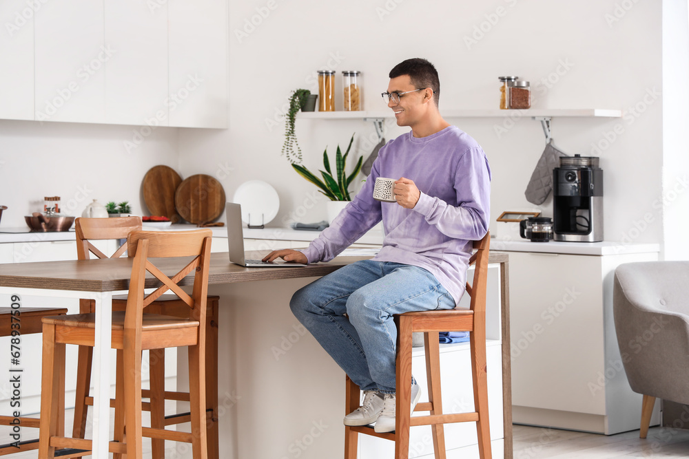 Handsome young man with cup of coffee using laptop while sitting in kitchen at home
