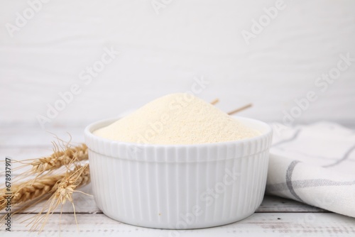 Uncooked organic semolina in bowl and spikelets on white wooden table, closeup