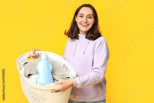 Young woman with laundry basket and bottle of detergent on yellow background