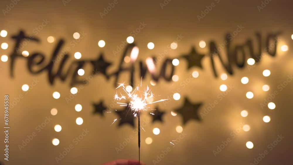 Hand holding a sparkling lit candle in celebration of the New Year, with a blurred banner in the background reading 'Happy New Year' in Portuguese.