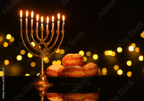 Menorah with burning candles and donuts for Hanukkah celebration on dark table against blurred lights photo