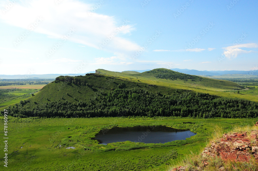 A small beautiful lake at the foot of a high hill of an unusual shape under a warm summer sky.