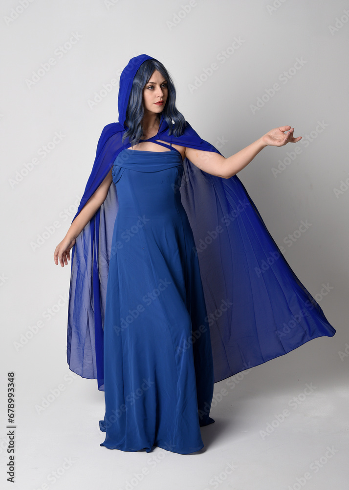 Naklejka premium Full length portrait of beautiful female model wearing elegant fantasy blue ball gown and flowing cape with hood. Standing pose, with gestural arms reaching out . Isolated on white studio background.