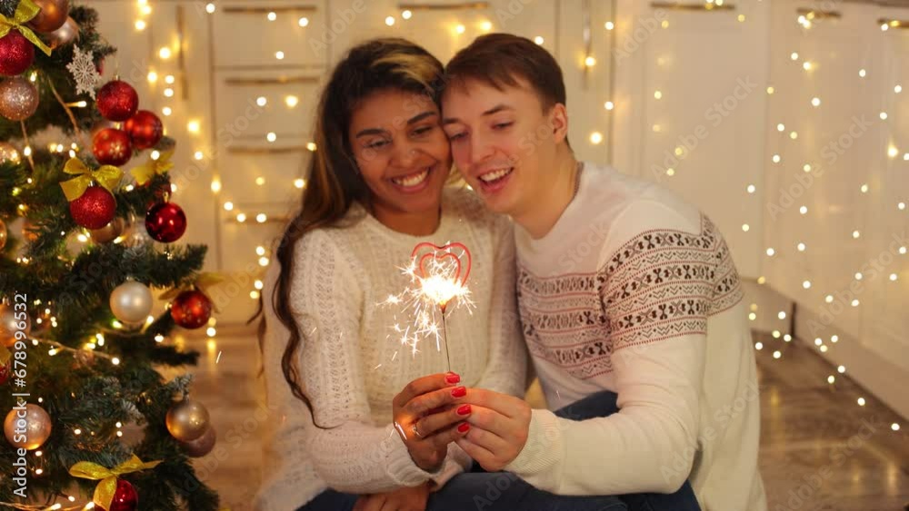 A couple in love creates a festive mood for themselves. The couple lights sparklers and spend time together on Christmas Eve. Two near the festively decorated Christmas tree in the house. A couple in love creates a festive mood for themselves. The couple lights sparklers and spend time together on Christmas Eve. Two near the festively decorated Christmas tree in the house.
