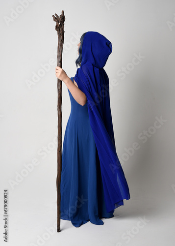 Full length portrait of beautiful female model wearing elegant fantasy blue ball gown, flowing cape with hood.Standing pose walking away, holding a wooden wizard staff. Isolated on white studio backgr