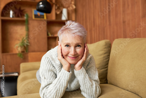 Portrait of lovely elderly female in warm jumper lying on brown couch leaning head on palms, having rest on cold winter day, relaxing, enjoying holidays, looking at camera with smile